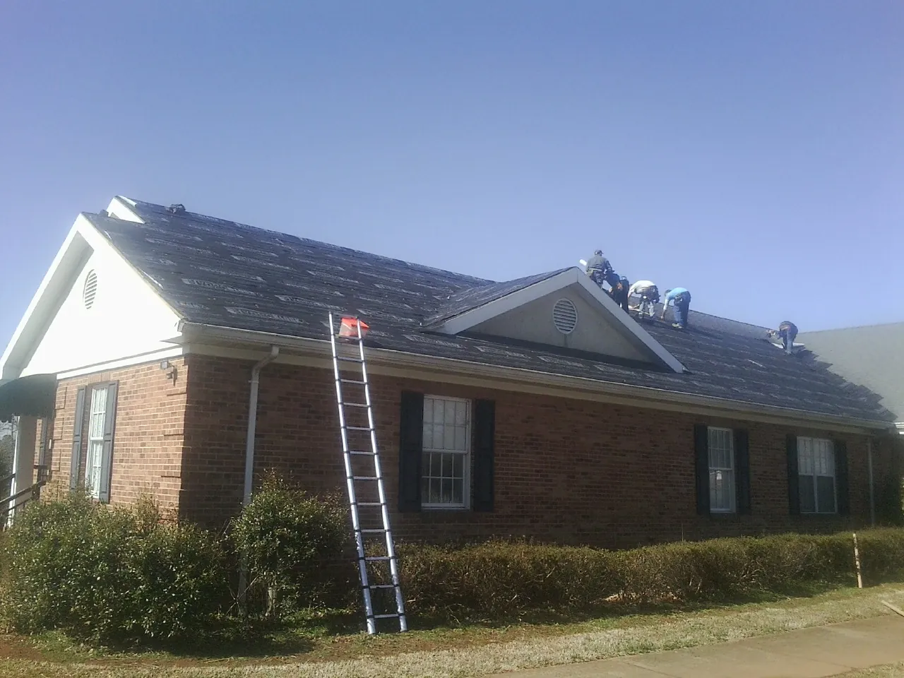 Crew tearing shingles off a brick ranch roof with a ladder set — Emergency Roof Repair work by our crew in West Edmeston
