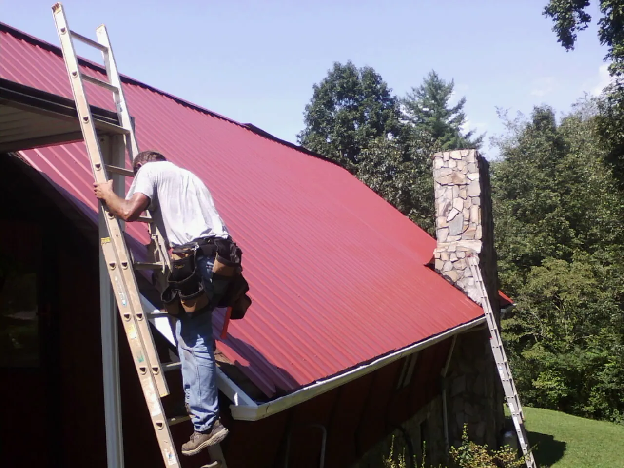 Roofer on a ladder beside a red metal roof and stone chimney — Soffit & Fascia Repair by Roofing Brothers in West Edmeston